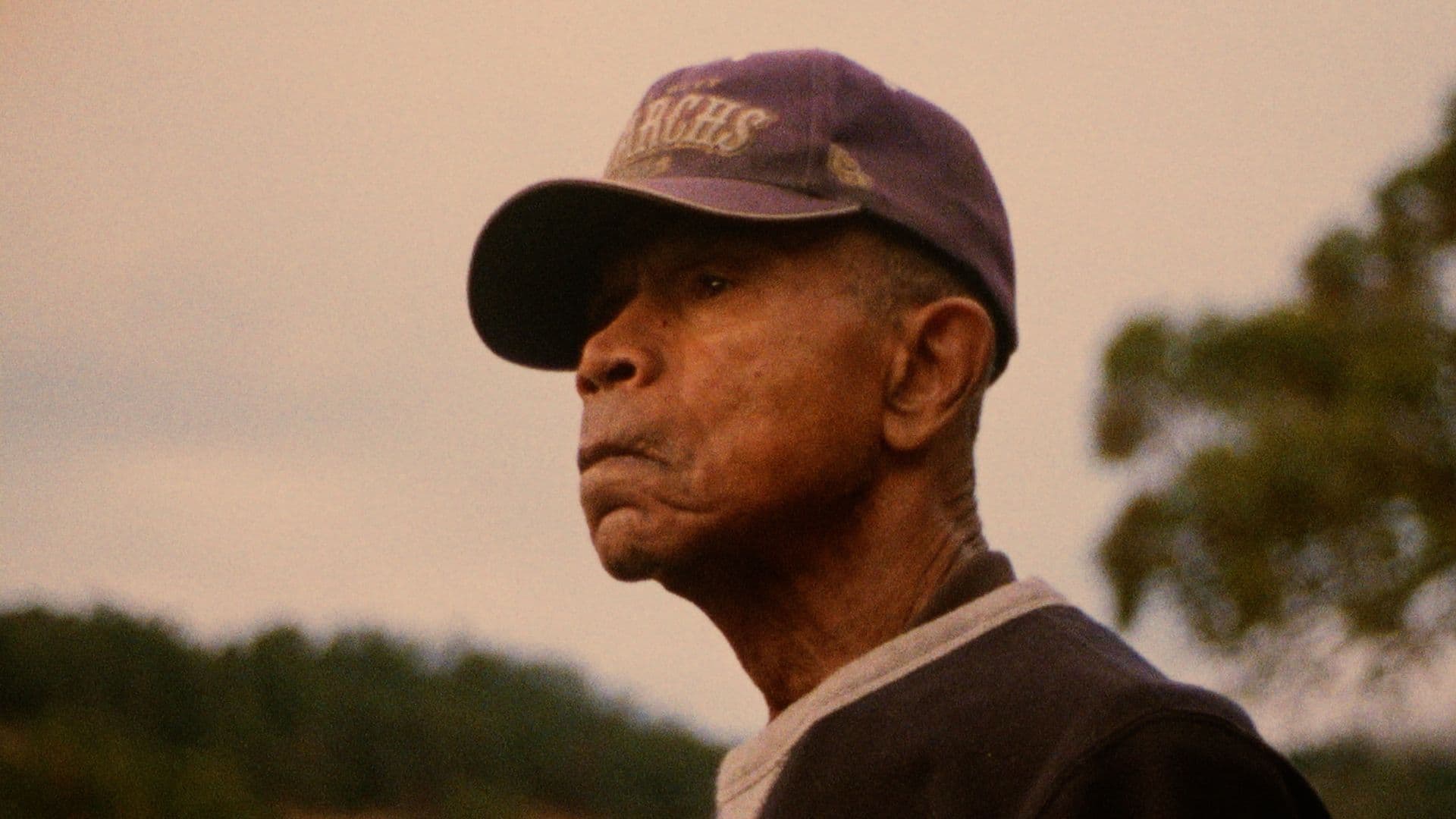 A film still from Buckskin shows an older Black man wearing a purple baseball cap looking off into the distance.