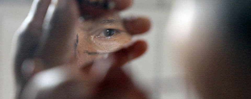 A film still from Charip: Lightning in the River shows an older Wampís man holding a mirror shard that reflects his eye.