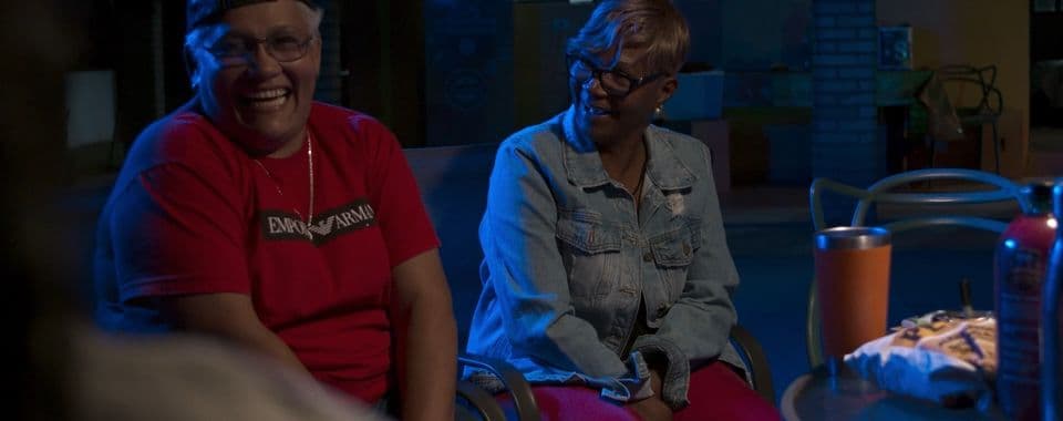 A film still from Communion shows two older Black woman sitting around a round table and smiling. The woman on the left wears a black cap and red shirt and the woman on the right is wearing a denim jacket and pink pants. The woman on the right is looking at the woman on the left.