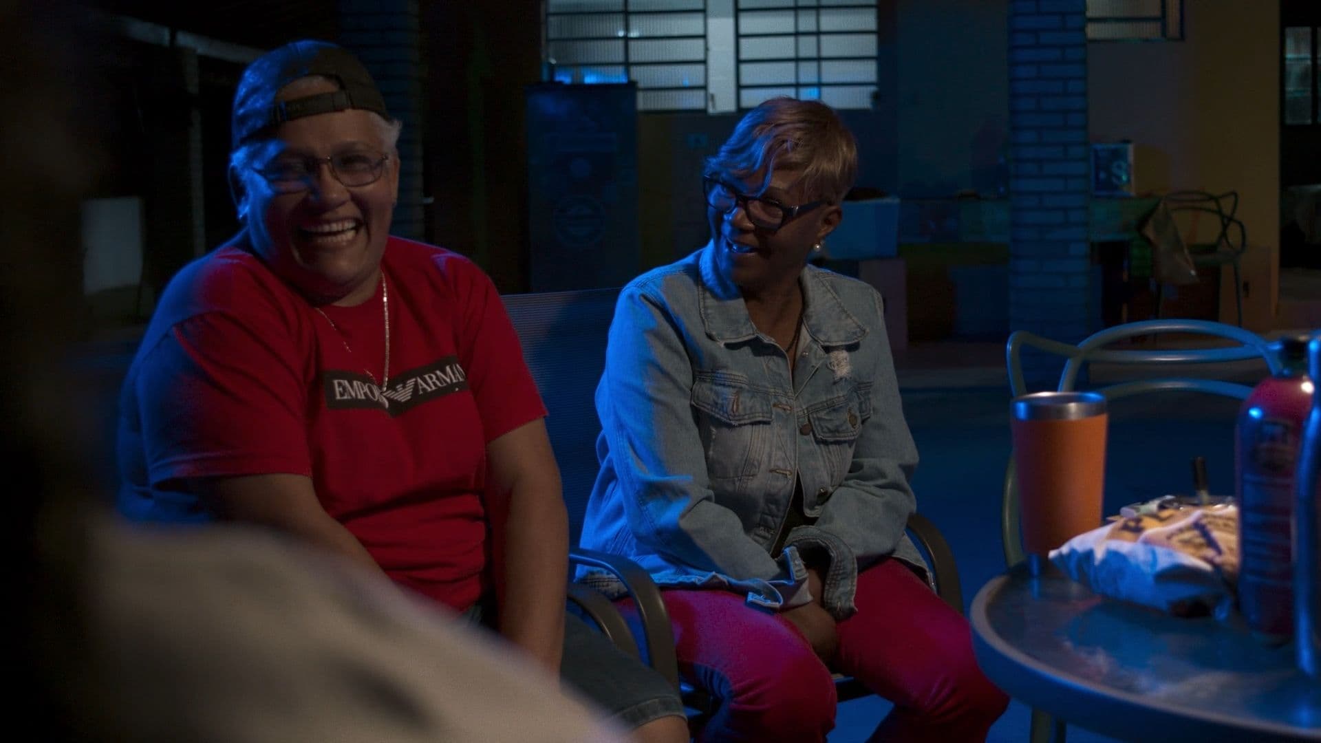 A film still from Communion shows two older Black woman sitting around a round table and smiling. The woman on the left wears a black cap and red shirt and the woman on the right is wearing a denim jacket and pink pants. The woman on the right is looking at the woman on the left.