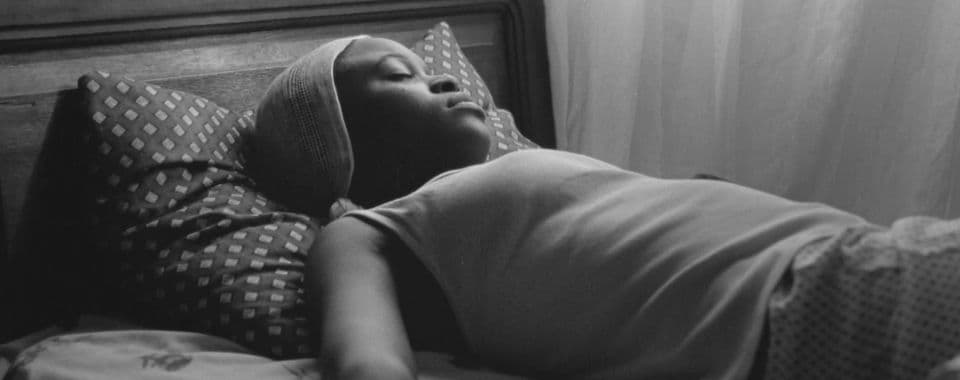 A black-and-white film still from Don’t wake the sleeping child shows a young Black girl fast asleep on her bed. She’s sleeping over the covers on a diamond-patterned pillow, facing a white curtain.