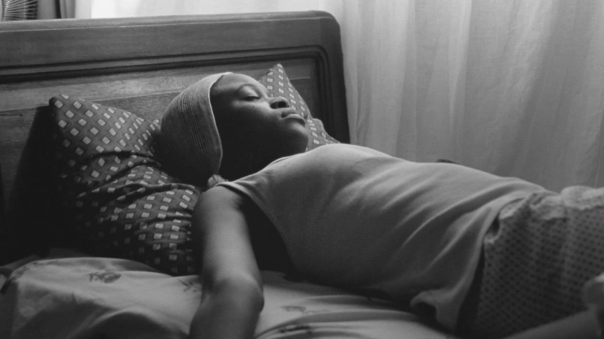 A black-and-white film still from Don’t wake the sleeping child shows a young Black girl fast asleep on her bed. She’s sleeping over the covers on a diamond-patterned pillow, facing a white curtain.