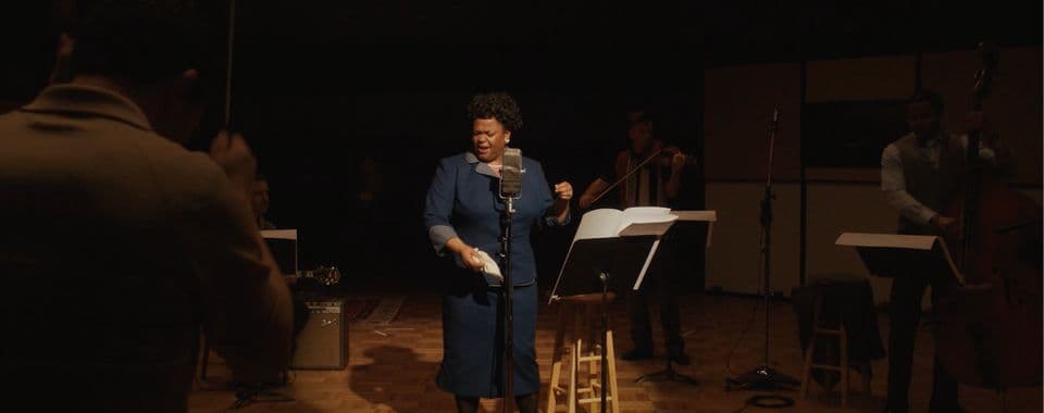 A film still from Ella shows a Black woman in a blue dress singing in a recording studio. She stands behind a microphone and next to a music stand and is lit by a spotlight. A bassist, violinist, and guitarist play around her and a conductor stands in the foreground.