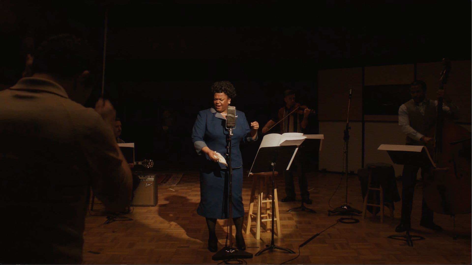 A film still from Ella shows a Black woman in a blue dress singing in a recording studio. She stands behind a microphone and next to a music stand and is lit by a spotlight. A bassist, violinist, and guitarist play around her and a conductor stands in the foreground.