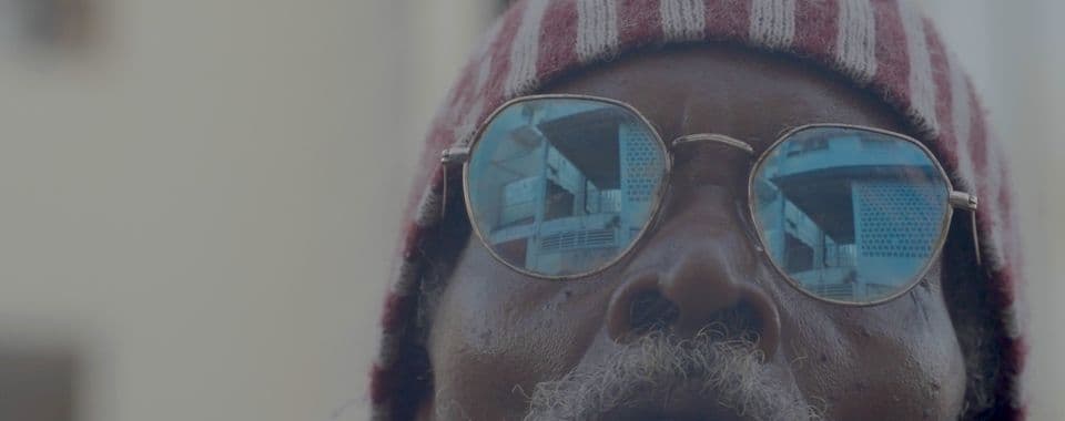 A film still from Enjoy the tropix and have a banana ! shows a close-up of an older Black man’s face. He has a gray beard and is wearing blue sunglasses and a red-and-white hood. In his sunglasses, there’s the reflection of older buildings.