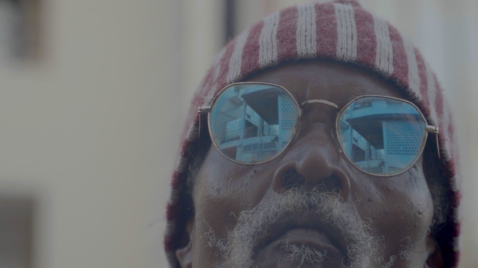 A film still from Enjoy the tropix and have a banana ! shows a close-up of an older Black man’s face. He has a gray beard and is wearing blue sunglasses and a red-and-white hood. In his sunglasses, there’s the reflection of older buildings.