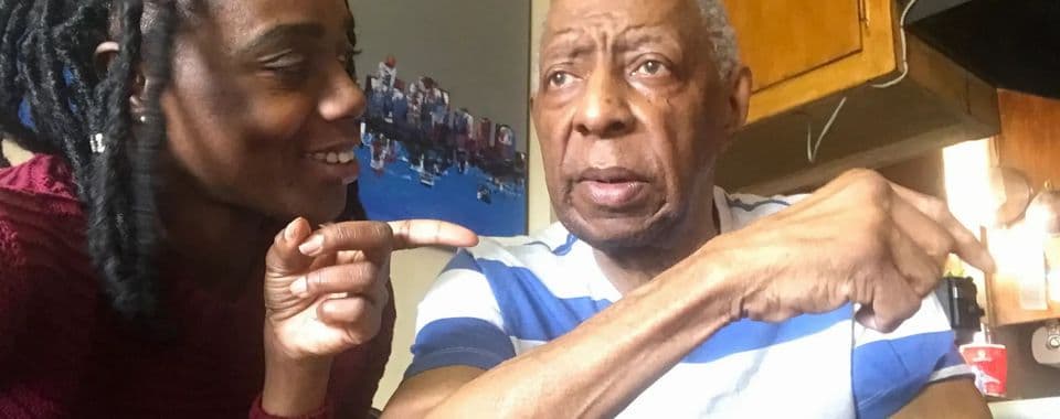 A film still from Finding Your Laughter shows a Black woman smiling and pointing toward an older Black man with short gray hair. The woman is wearing a red sweater and the man is wearing a blue-and-white shirt.
