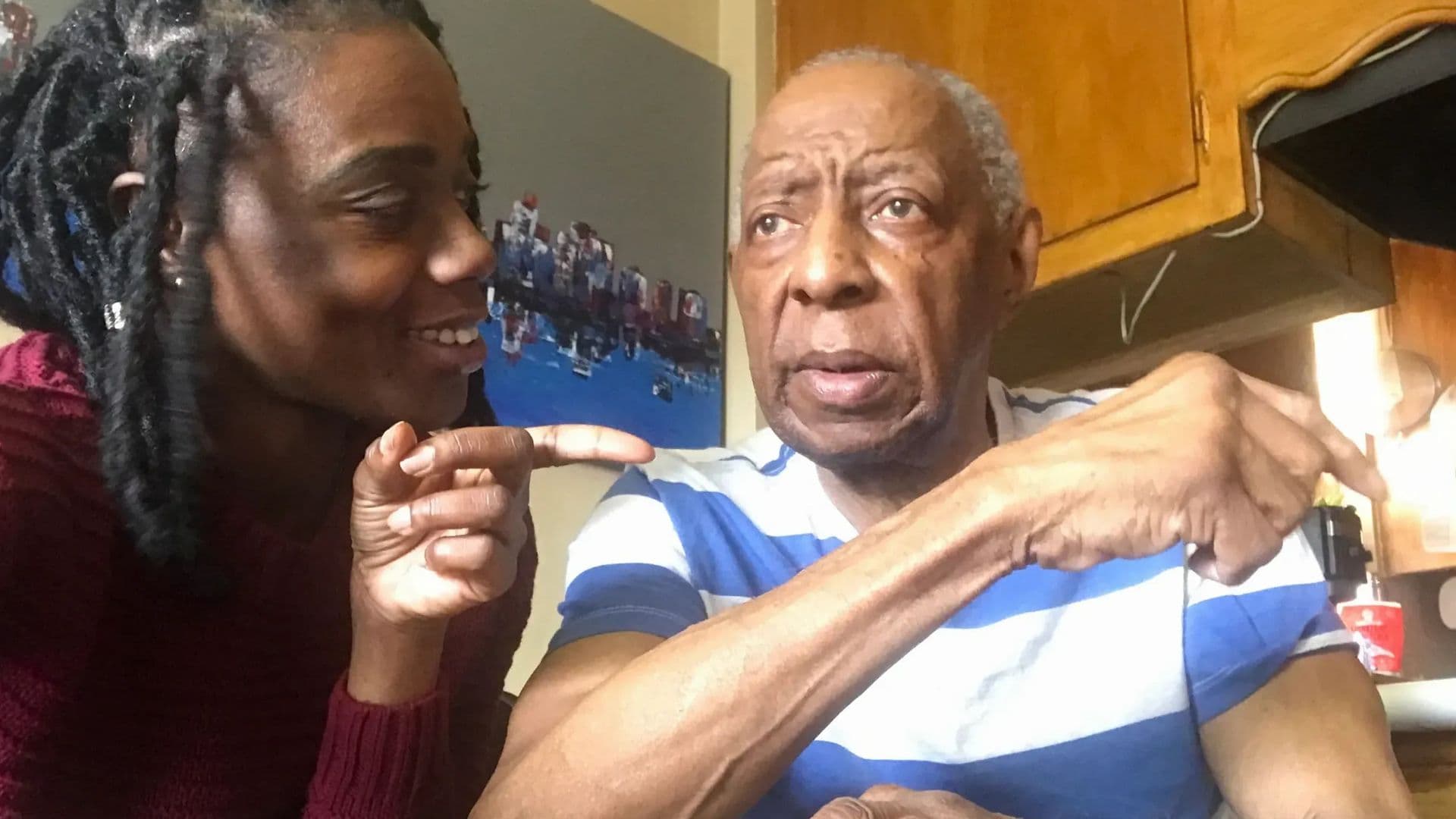 A film still from Finding Your Laughter shows a Black woman smiling and pointing toward an older Black man with short gray hair. The woman is wearing a red sweater and the man is wearing a blue-and-white shirt.