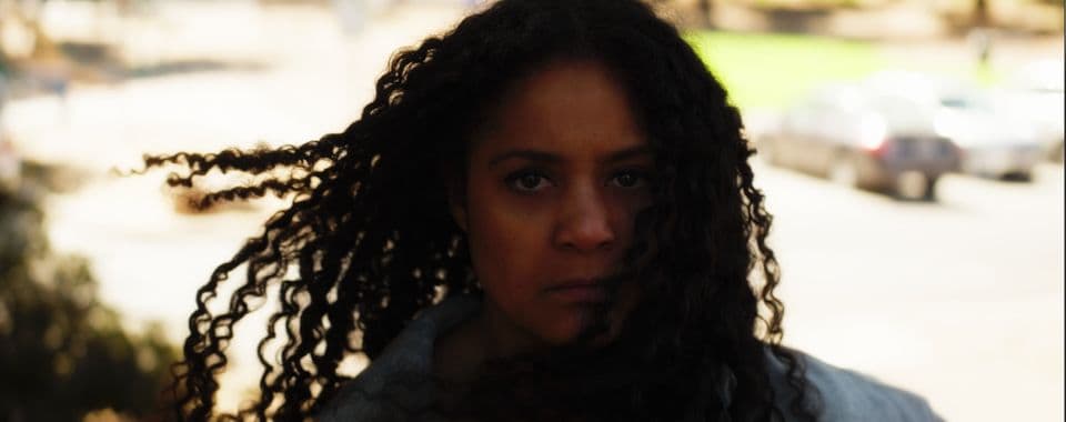 A film still from Fists of Mothers shows a young Black woman with long hair looking directly into the camera with a serious expression. She is standing outside on a sunny street. Her hair is being blown by the wind.
