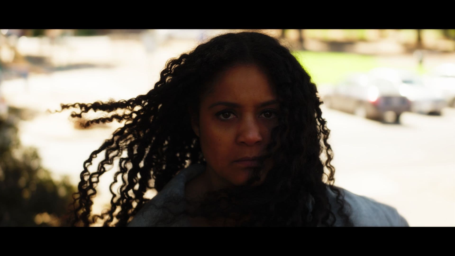 A film still from Fists of Mothers shows a young Black woman with long hair looking directly into the camera with a serious expression. She is standing outside on a sunny street. Her hair is being blown by the wind.