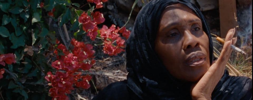 A film still from For Peace of Mind shows a Black woman in a black headscarf sitting in front of a stone wall with red flowers. She is propping her chin on her left hand and looking off into the distance.