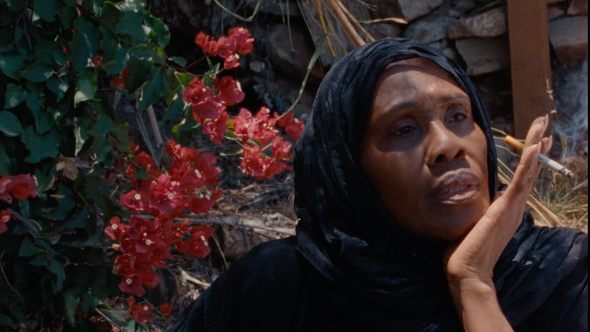 A film still from For Peace of Mind shows a Black woman in a black headscarf sitting in front of a stone wall with red flowers. She is propping her chin on her left hand and looking off into the distance.
