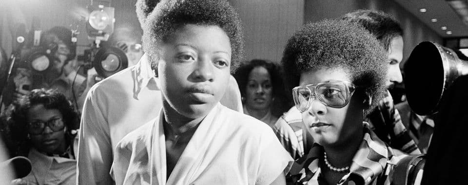 A black-and-white film still from Free Joan Little shows Little, a young Black woman in a white top, standing beside another Black woman in glasses. They are standing in a hallway, surrounded by journalists and photographers.