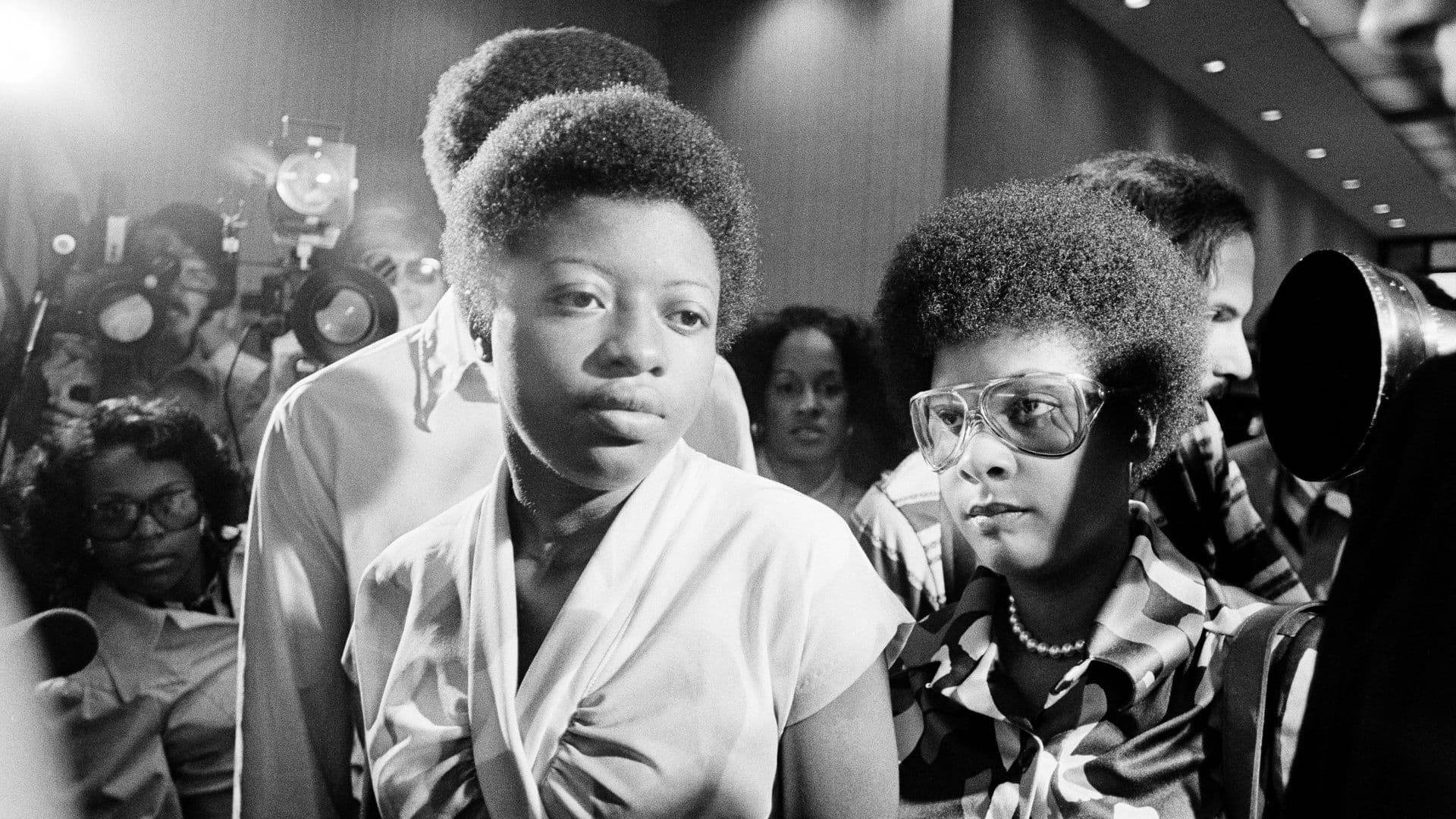 A black-and-white film still from Free Joan Little shows Little, a young Black woman in a white top, standing beside another Black woman in glasses. They are standing in a hallway, surrounded by journalists and photographers.