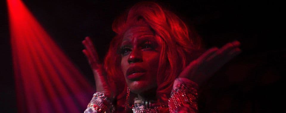 A film still from Glass Bricks shows a Black drag queen performing in a dark room with a red spotlight.
