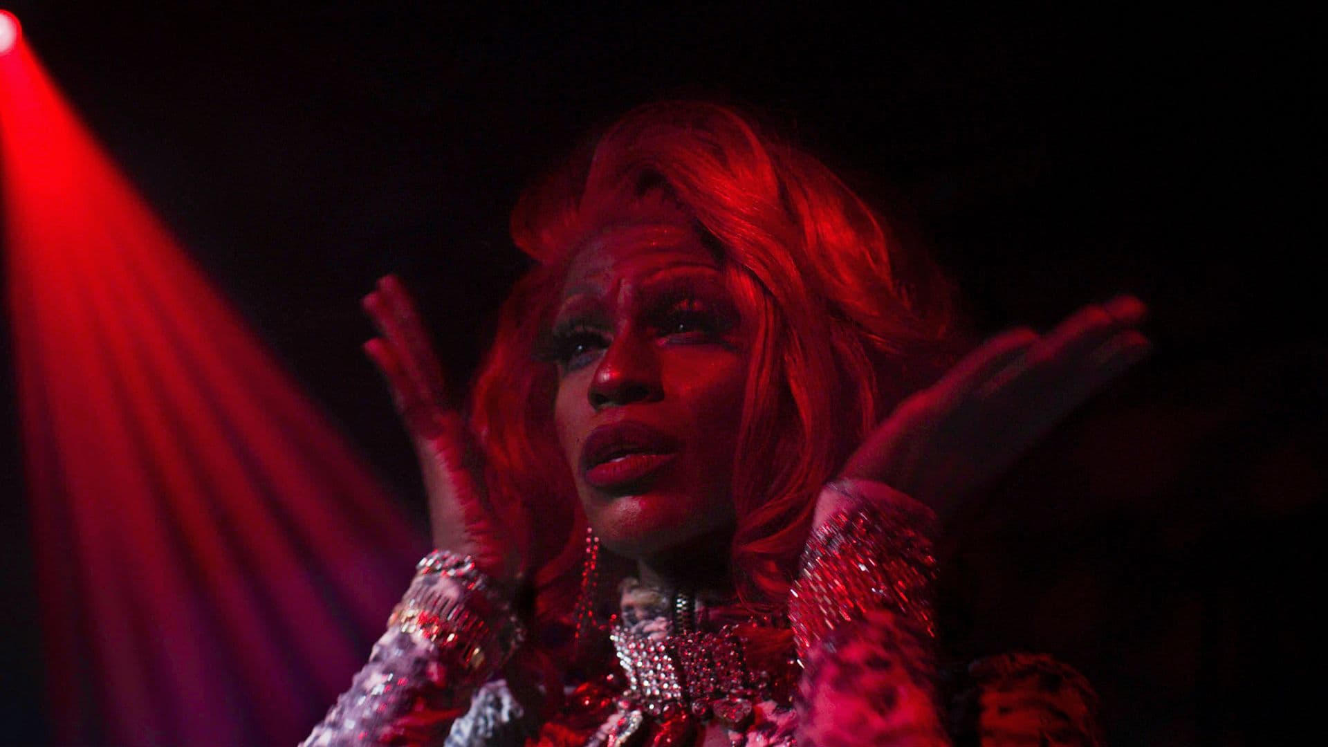 A film still from Glass Bricks shows a Black drag queen performing in a dark room with a red spotlight.