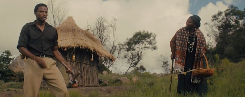 A film still from God Sleeps on Sundays shows a young Black man and an older Black woman standing in front of a wooden hut. The man holds a hammer and looks into the distance. The woman holds a cane and a basket and is speaking to the man.