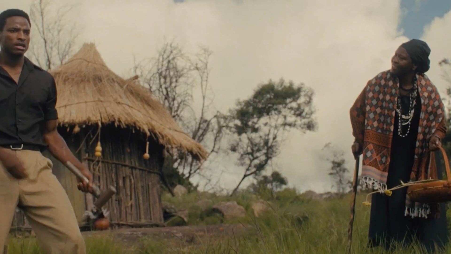 A film still from God Sleeps on Sundays shows a young Black man and an older Black woman standing in front of a wooden hut. The man holds a hammer and looks into the distance. The woman holds a cane and a basket and is speaking to the man.