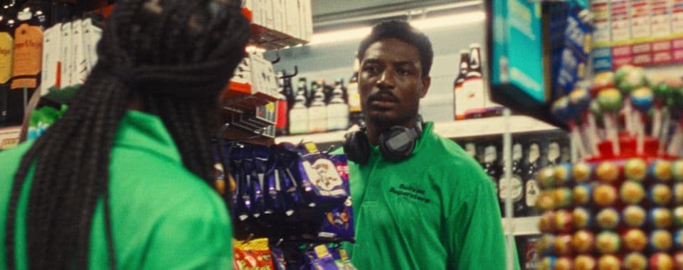 A film still from How To Hide It shows two Black people facing each other inside a convenience store. They are both wearing bright green polo shirts labeled: “Sullivan Superstore.” The person on the left has box braids falling down their back. The young Black man on the right is facing the other person and wears headphones around his neck. The store has shelves stocked with bottles, candies, and more.