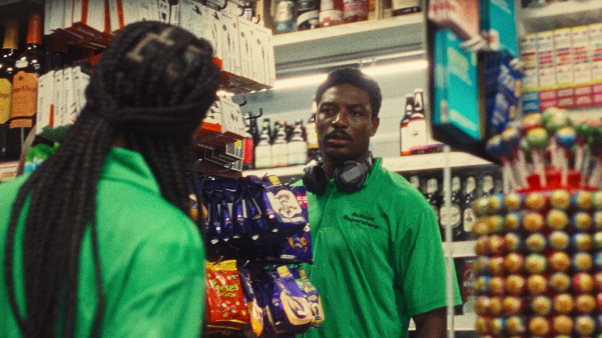 A film still from How To Hide It shows two Black people facing each other inside a convenience store. They are both wearing bright green polo shirts labeled: “Sullivan Superstore.” The person on the left has box braids falling down their back. The young Black man on the right is facing the other person and wears headphones around his neck. The store has shelves stocked with bottles, candies, and more.