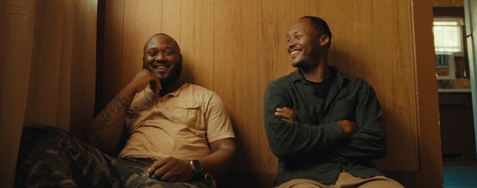 A film still from I Got My Brother shows two Black men sitting on the floor against a wood wall in a house. The man on the left is wearing a beige shirt and camo pants. He has a tattoo on his right arm. The man on the right is wearing a green shirt and khaki pants. He’s looking at the man on the left, and the two are both smiling.