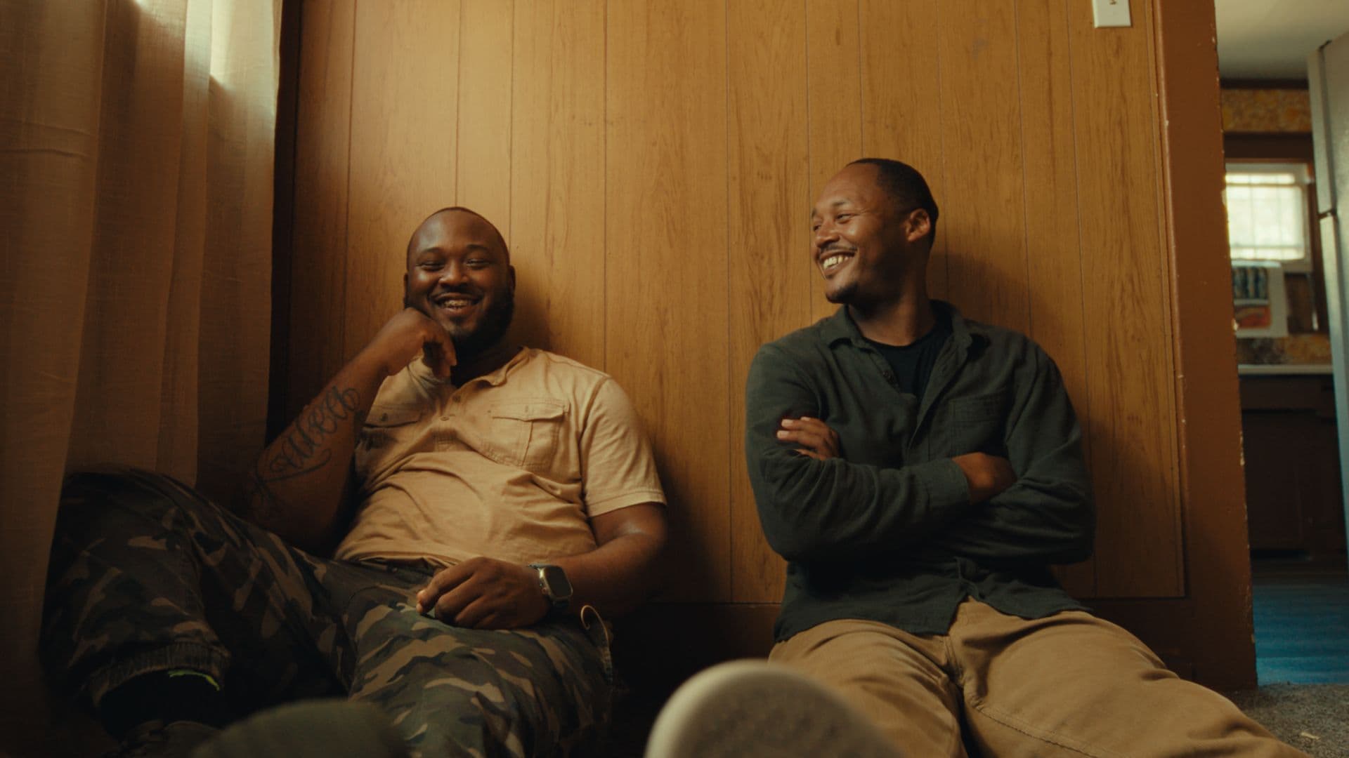 A film still from I Got My Brother shows two Black men sitting on the floor against a wood wall in a house. The man on the left is wearing a beige shirt and camo pants. He has a tattoo on his right arm. The man on the right is wearing a green shirt and khaki pants. He’s looking at the man on the left, and the two are both smiling.