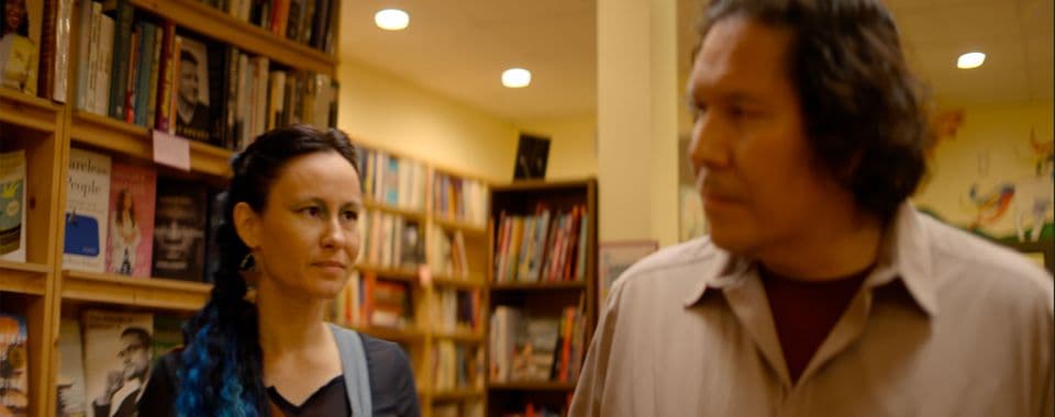 A film still from Ish Meets a Mermaid shows an Anishinaabe woman and an Ojibwe man standing in a bookstore. The woman has blue hair extensions and blue eyeshadow, and she is staring at the man.