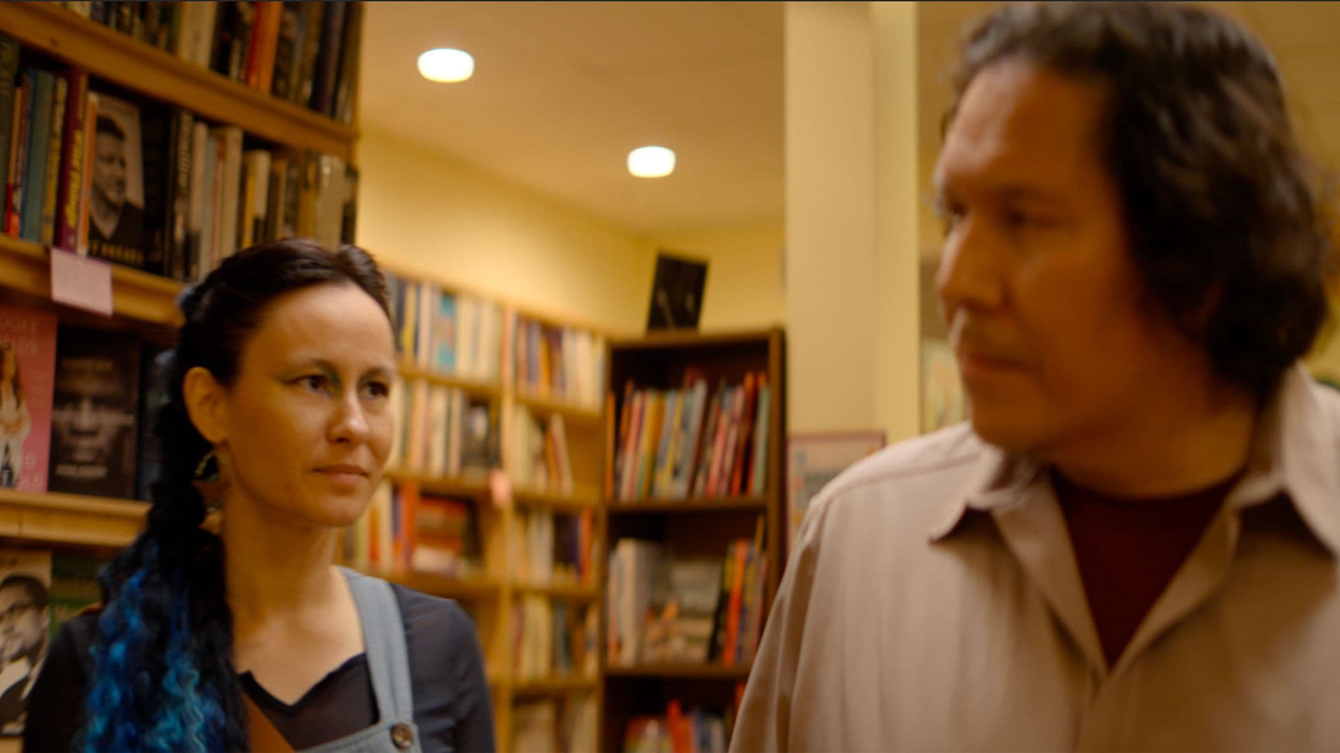 A film still from Ish Meets a Mermaid shows an Anishinaabe woman and an Ojibwe man standing in a bookstore. The woman has blue hair extensions and blue eyeshadow, and she is staring at the man.