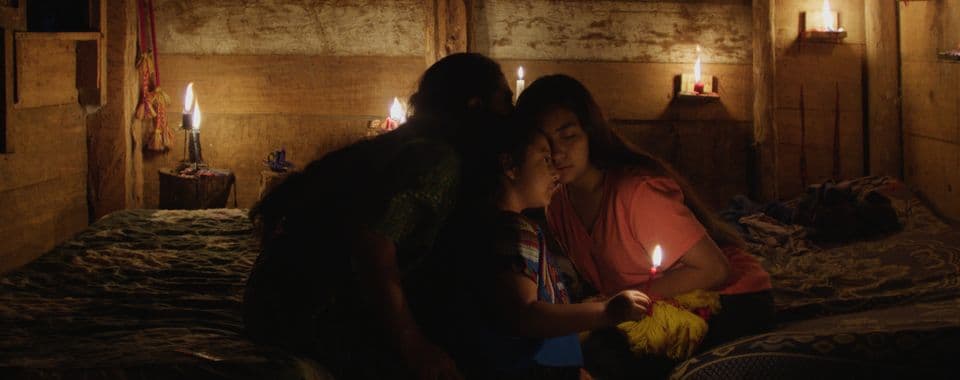 A film still from Ixquic shows two Indigenous children and an older Indigenous woman sitting on two beds in a wooden room. The room is illuminated by candles. The younger child is holding a candle while the older woman whispers to the other child, a young girl in a pink shirt.