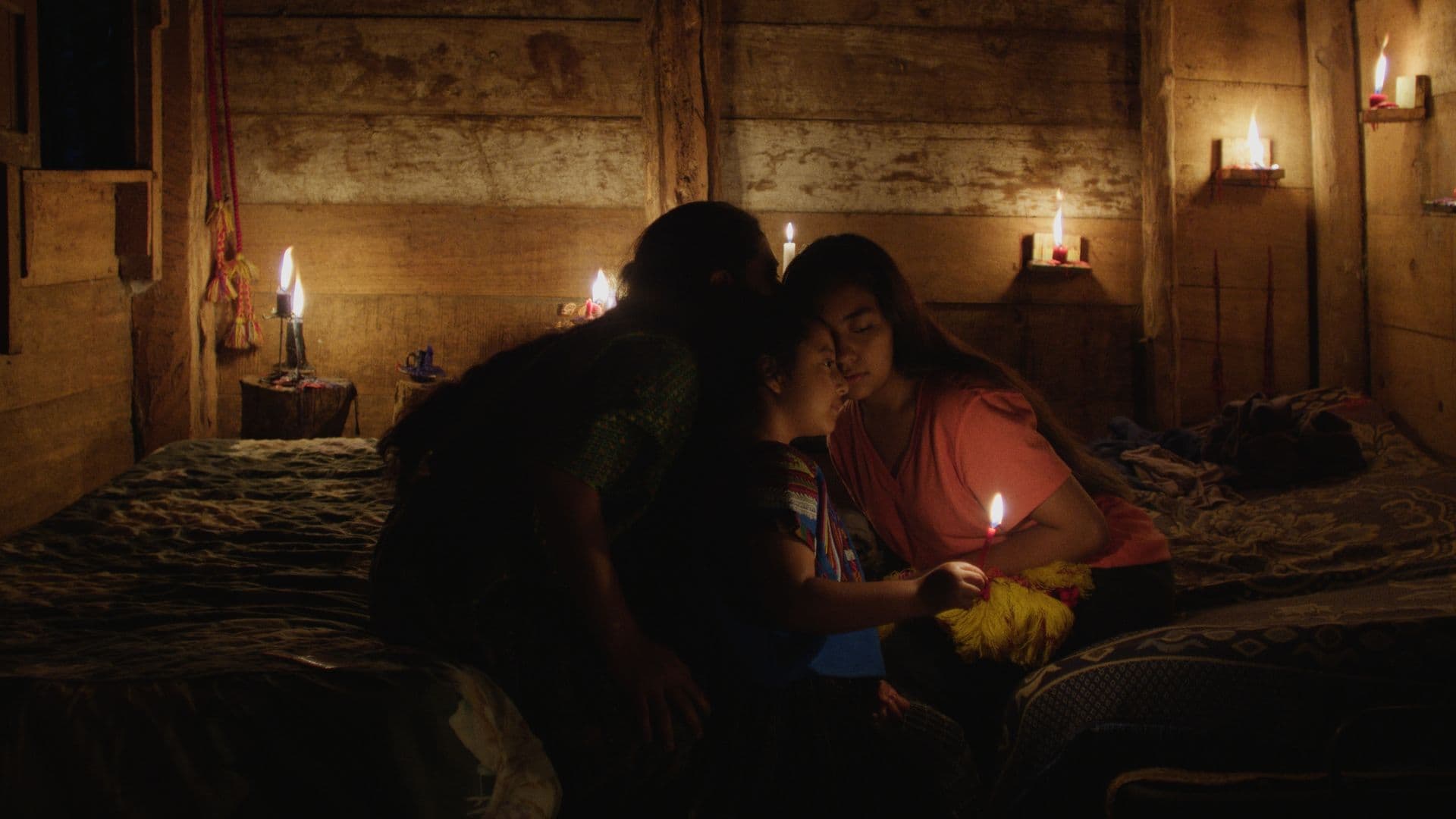 A film still from Ixquic shows two Indigenous children and an older Indigenous woman sitting on two beds in a wooden room. The room is illuminated by candles. The younger child is holding a candle while the older woman whispers to the other child, a young girl in a pink shirt.