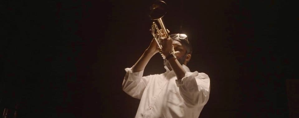 A film still from Lani B Supreme: Legacy shows a Black man in a white shirt playing a trumpet. The man is illuminated by a bright light but surrounded by darkness, as if performing on a stage.