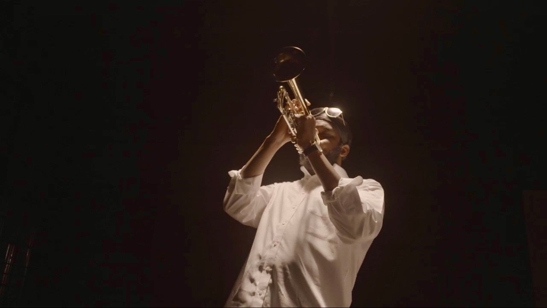 A film still from Lani B Supreme: Legacy shows a Black man in a white shirt playing a trumpet. The man is illuminated by a bright light but surrounded by darkness, as if performing on a stage.
