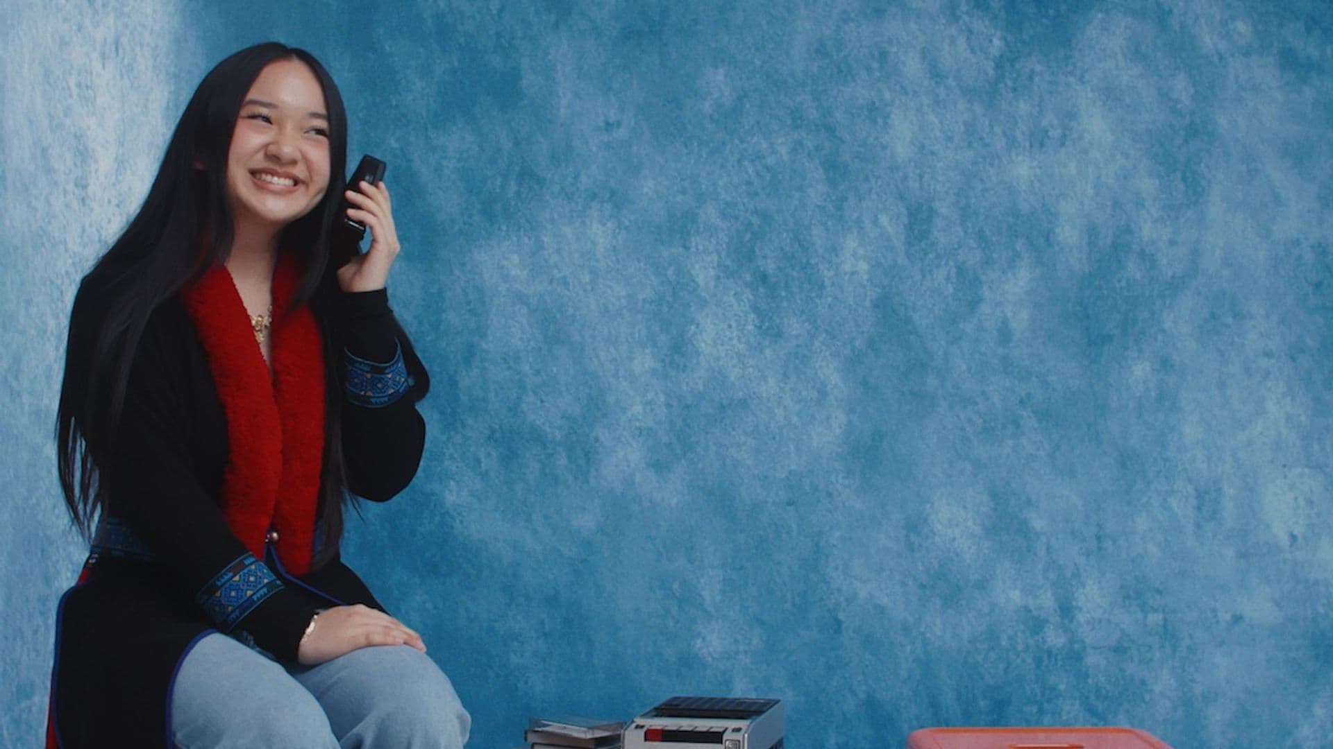 A film still from Muoz-Doic Mixtape shows a young Mien woman sitting on an orange stool in front of a blue backdrop. She is holding a black portable cassette player next to her ear and smiling. Next to her, there’s a wooden round table and another orange stool.