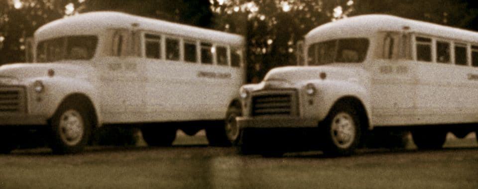 A sepia-toned still from National Seating shows two school buses parked side-by-side on a driveway, surrounded by grass and trees.
