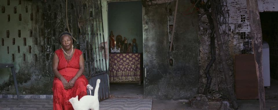 A film still from No Sunshine In Here shows an older Black woman in a red dress sitting in front of a run-down building. Behind the woman, there’s an open door revealing a table with statues and a candle. A white cat paces at the woman’s feet, looking up at her.