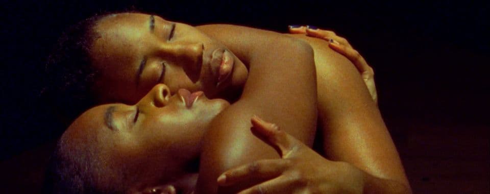A film still from Nwanne M Nwaanyị shows two Black people wrapped in a close embrace, facing up. Their face and body shine in the yellow light while their surroundings are dark. Their eyes are closed and their expressions are peaceful.