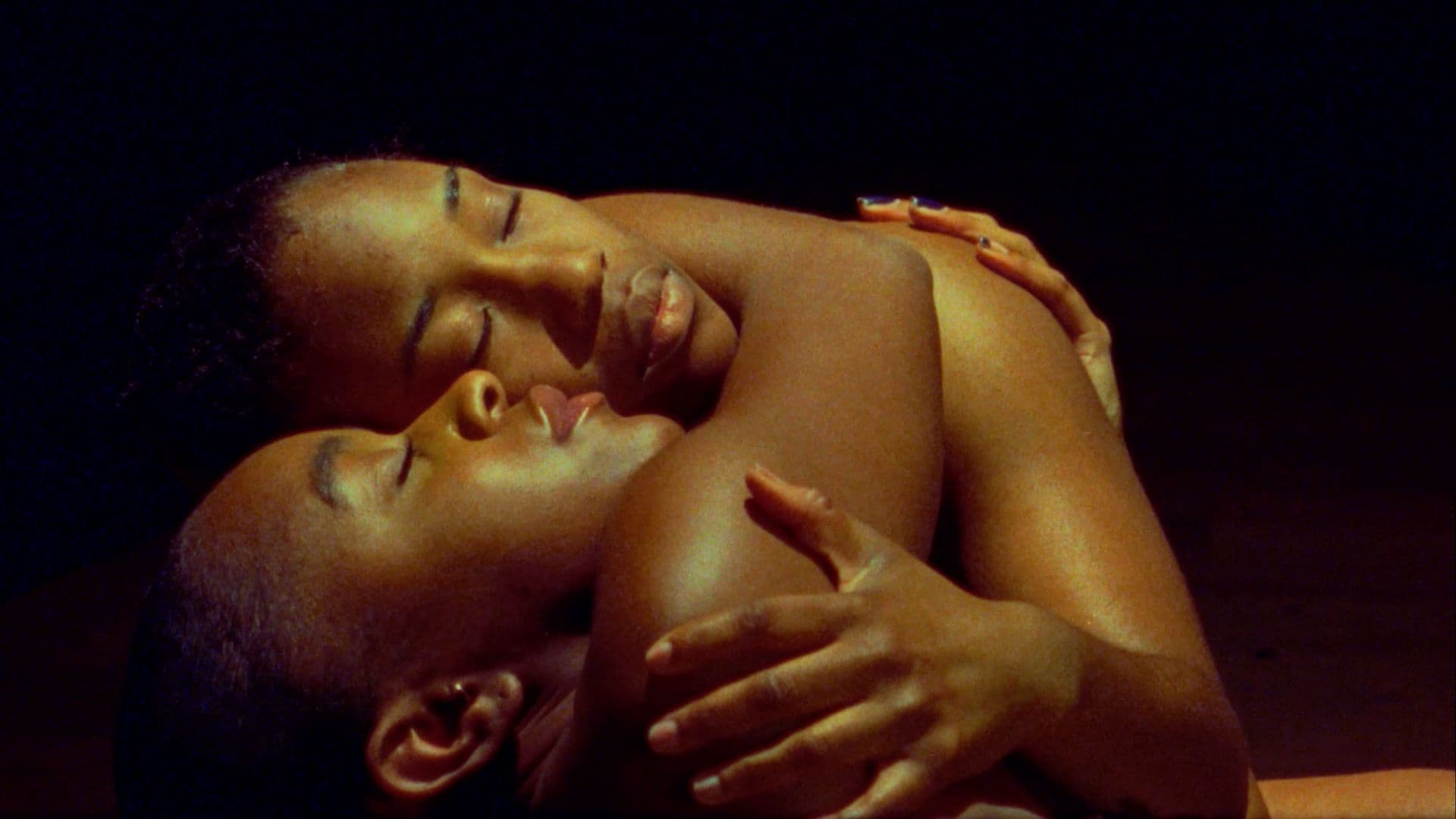 A film still from Nwanne M Nwaanyị shows two Black people wrapped in a close embrace, facing up. Their face and body shine in the yellow light while their surroundings are dark. Their eyes are closed and their expressions are peaceful.