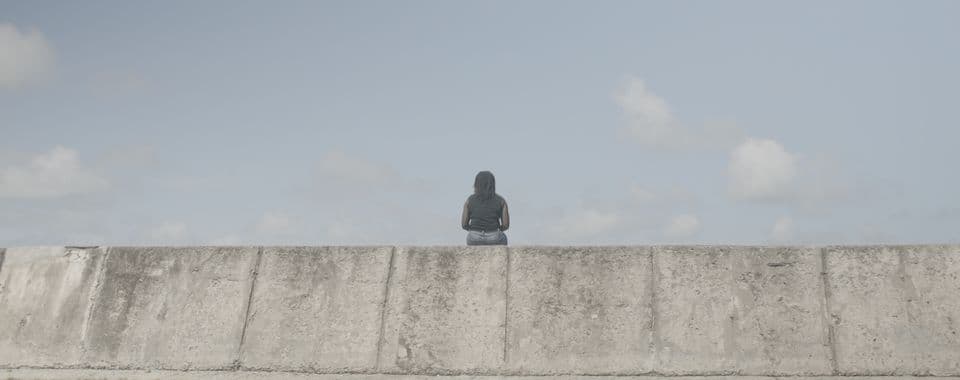 A film still from Old Man River shows a Black woman sitting on a sea wall, facing away from the camera. She’s wearing a black top and jeans. The sky in front of her is blue, with a few clouds.