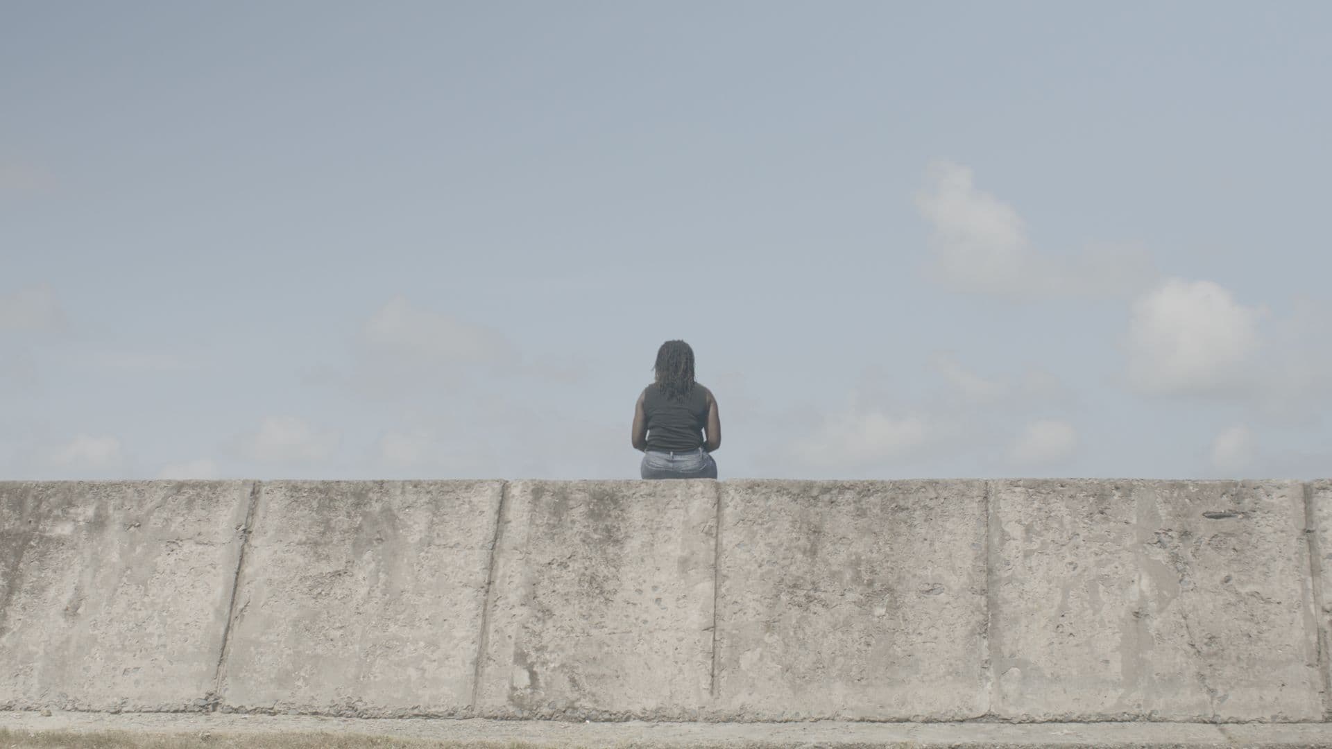 A film still from Old Man River shows a Black woman sitting on a sea wall, facing away from the camera. She’s wearing a black top and jeans. The sky in front of her is blue, with a few clouds.