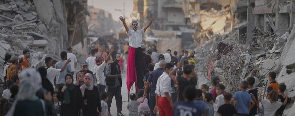 A film still from One More Show shows a Palestinian man on stilts raising his arms above a crowd of people walking in the ruins of a neighborhood. The man is wearing clown makeup and red-and-black pants. Some people in the crowd are watching him while others walk past.