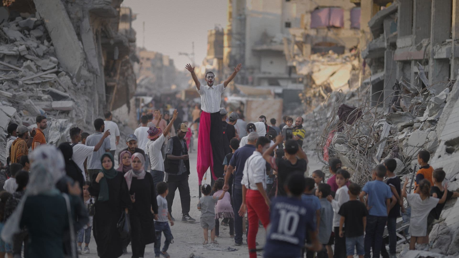 A film still from One More Show shows a Palestinian man on stilts raising his arms above a crowd of people walking in the ruins of a neighborhood. The man is wearing clown makeup and red-and-black pants. Some people in the crowd are watching him while others walk past.