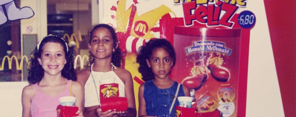 A film still from Olinda’s Golden Arches shows three children standing side-by-side inside a McDonald's, looking forward and holding up red Happy Meal trays with drinks and food boxes. From left to right, the first child smiles brightly, another poses confidently, and the third looks at the camera with a more serious expression. Behind them, a large promotional display shows Ronald McDonald.