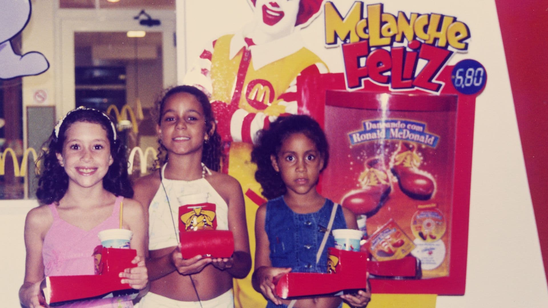 A film still from Olinda’s Golden Arches shows three children standing side-by-side inside a McDonald's, looking forward and holding up red Happy Meal trays with drinks and food boxes. From left to right, the first child smiles brightly, another poses confidently, and the third looks at the camera with a more serious expression. Behind them, a large promotional display shows Ronald McDonald.