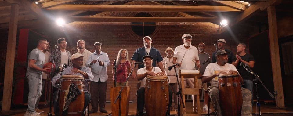 A film still from Philly Rumba shows a group of majority Black and Latin American men at a recording studio. There are three older Black men sitting in the front, playing hand drums with microphone stands next to them. Behind them, the rest of the group is standing or clapping. The studio has wooden beams along the ceiling and a brick wall.