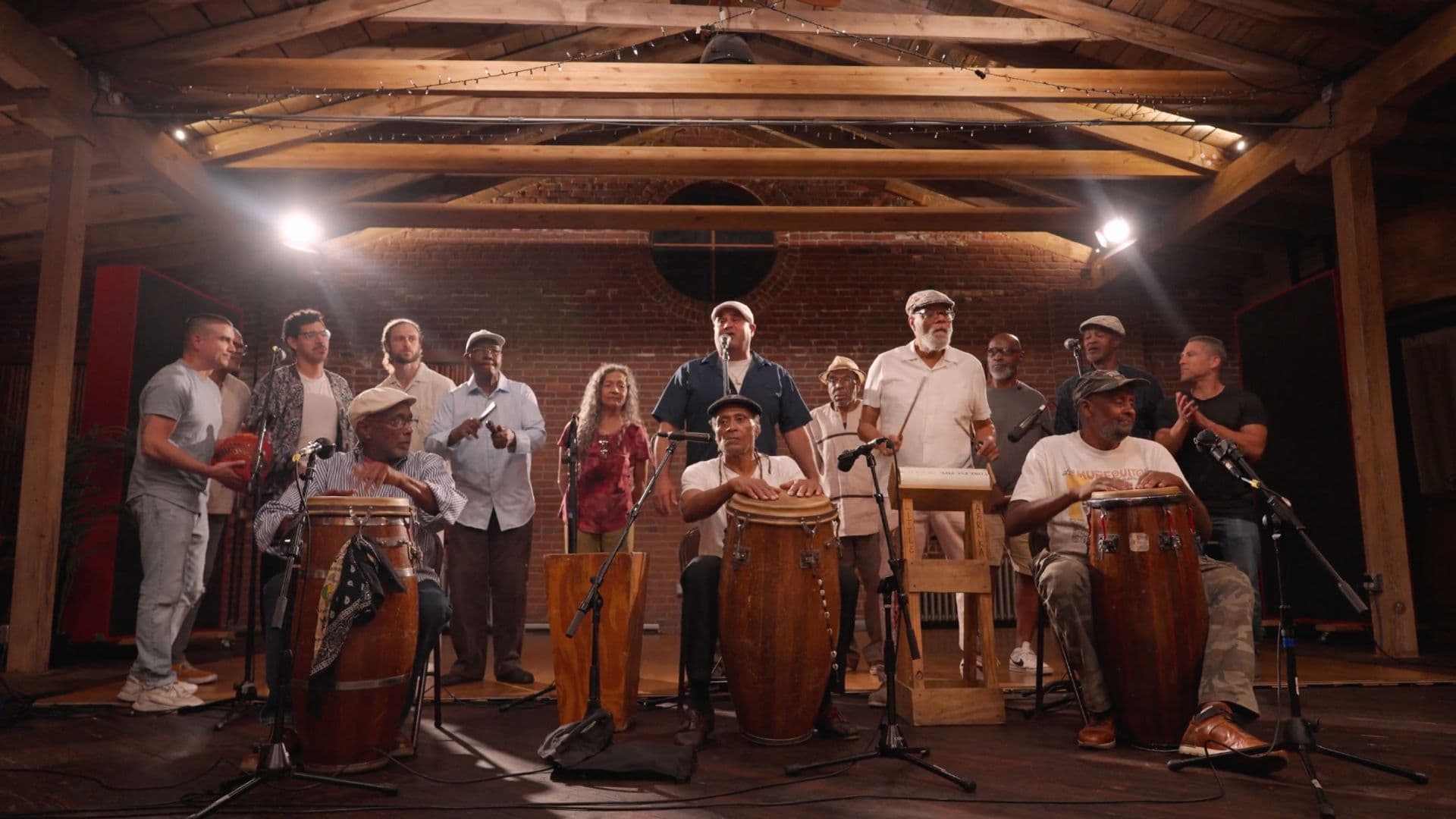 A film still from Philly Rumba shows a group of majority Black and Latin American men at a recording studio. There are three older Black men sitting in the front, playing hand drums with microphone stands next to them. Behind them, the rest of the group is standing or clapping. The studio has wooden beams along the ceiling and a brick wall.