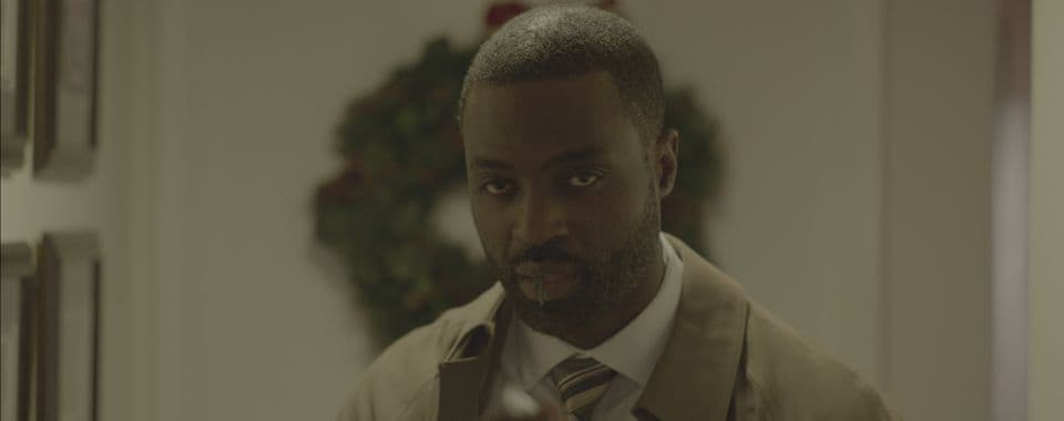 A film still from Potato Salad shows a Black man in a hallway looking forward. He is wearing a khaki trenchcoat, a white shirt, and a tie. From his mouth, hangs a sprig of pine. There are frames along the wall and a Christmas wreath hanging on the door behind him.