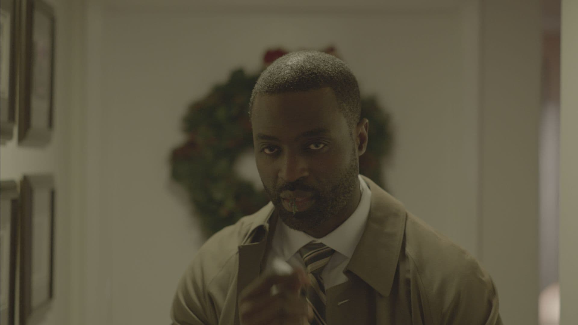 A film still from Potato Salad shows a Black man in a hallway looking forward. He is wearing a khaki trenchcoat, a white shirt, and a tie. From his mouth, hangs a sprig of pine. There are frames along the wall and a Christmas wreath hanging on the door behind him.