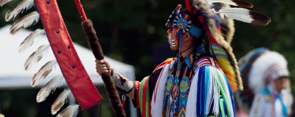 A film still from Powwow People shows a Native American man in full regalia holding an eagle staff. He holds the staff in his right hand and faces toward the left of the frame. The man’s regalia includes colorful decorative beadwork and a headdress. The eagle staff is red with white feathers coming off it.