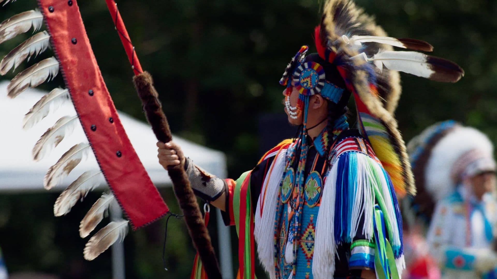 A film still from Powwow People shows a Native American man in full regalia holding an eagle staff. He holds the staff in his right hand and faces toward the left of the frame. The man’s regalia includes colorful decorative beadwork and a headdress. The eagle staff is red with white feathers coming off it.