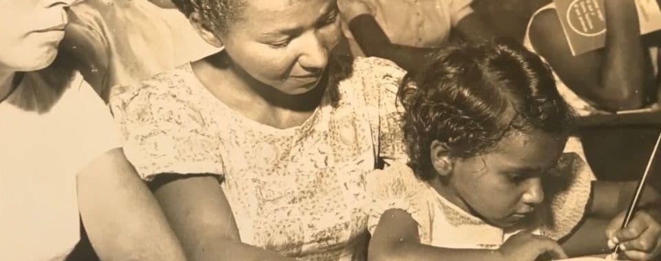 A sepia-toned film still from Reading the World shows a young Brazilian girl sitting on the lap of an older Brazilian woman as they look at a piece of paper on a desk. The girl is holding a pencil and writing on the paper. Around them are other women sitting at desks.